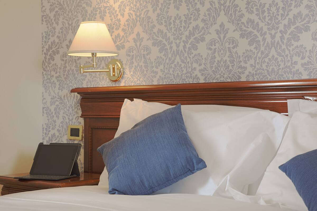 A neatly made bed with white and blue pillows, a laptop on a bedside table, and a wall-mounted lamp.