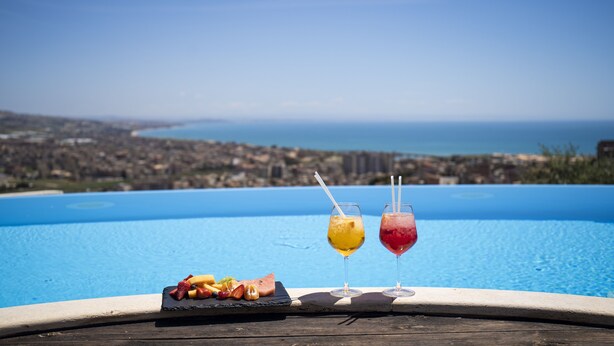 A poolside view with a wooden deck, two glasses of fruit juice, and a fruit platter. - Villa Giuliana Relais (Licata)