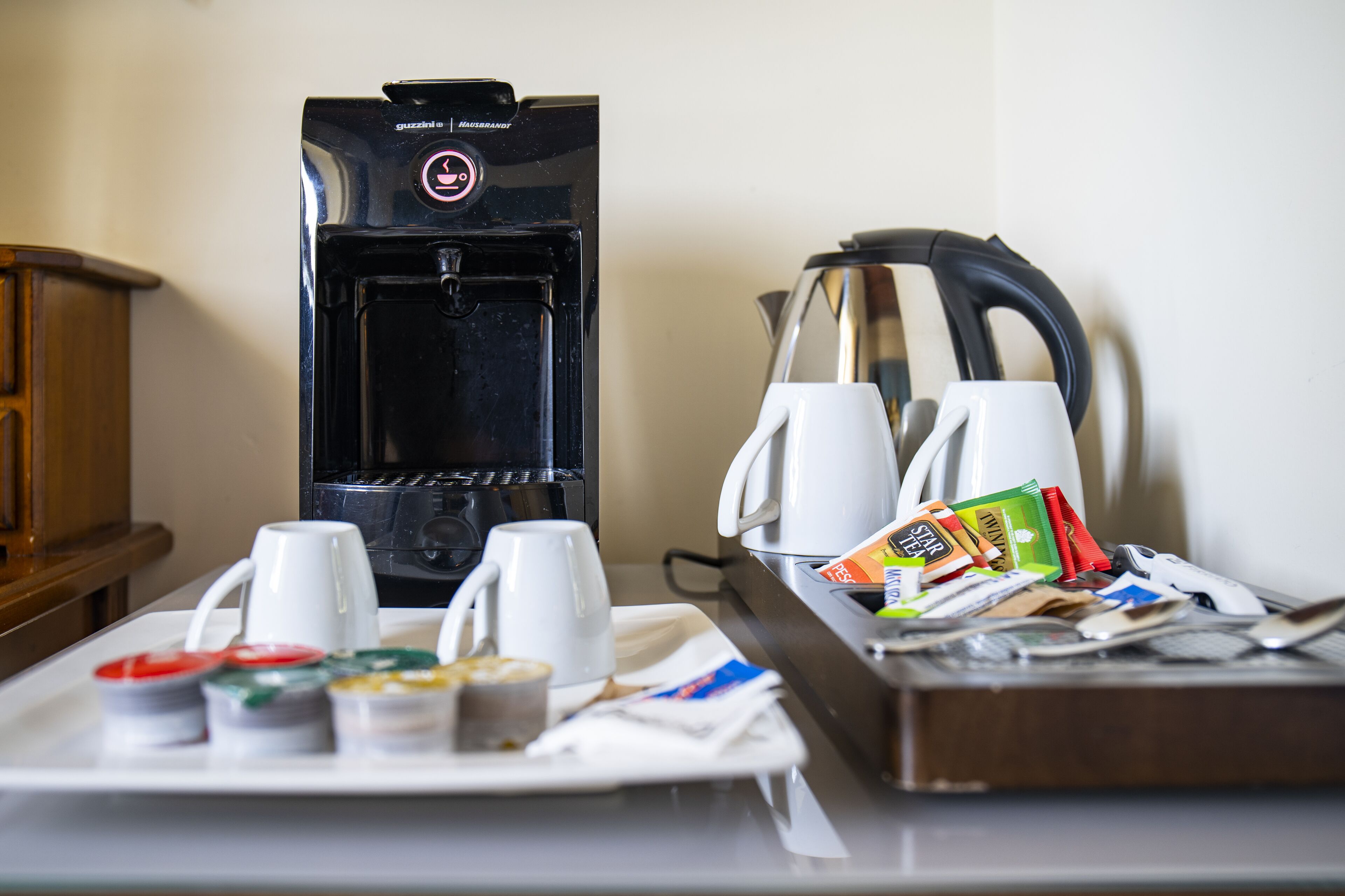 A coffee machine, two white cups, a tray with food items, and a kettle on a table.