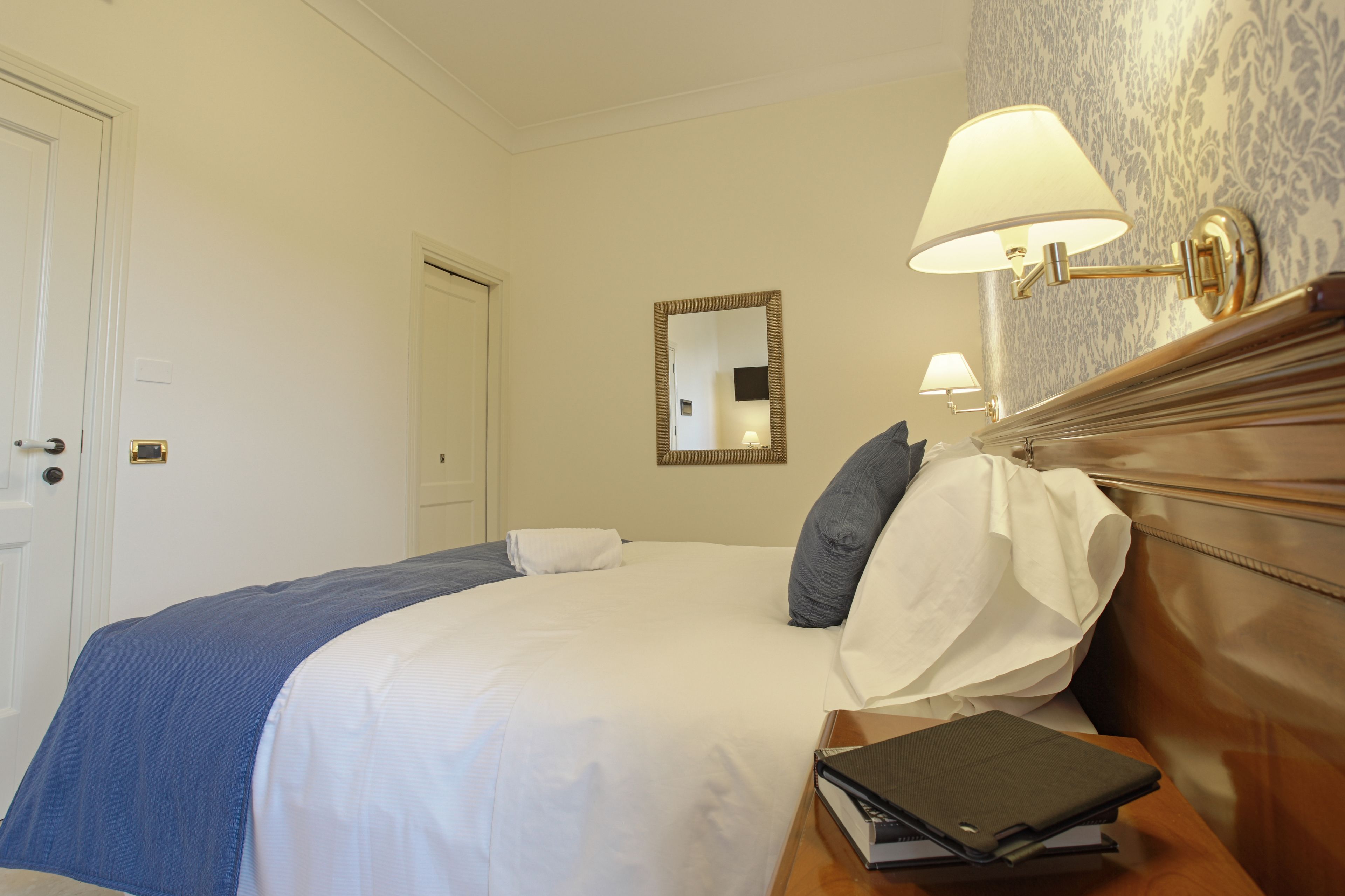 A neatly made bed with a blue blanket, a white pillow, and a grey pillow. There is a wooden nightstand with a book and a lamp.