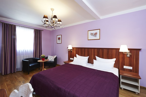 A hotel room with a large bed, a wooden headboard, two bedside tables with lamps, a small round table with a plant, a sofa, and a large window with curtains.