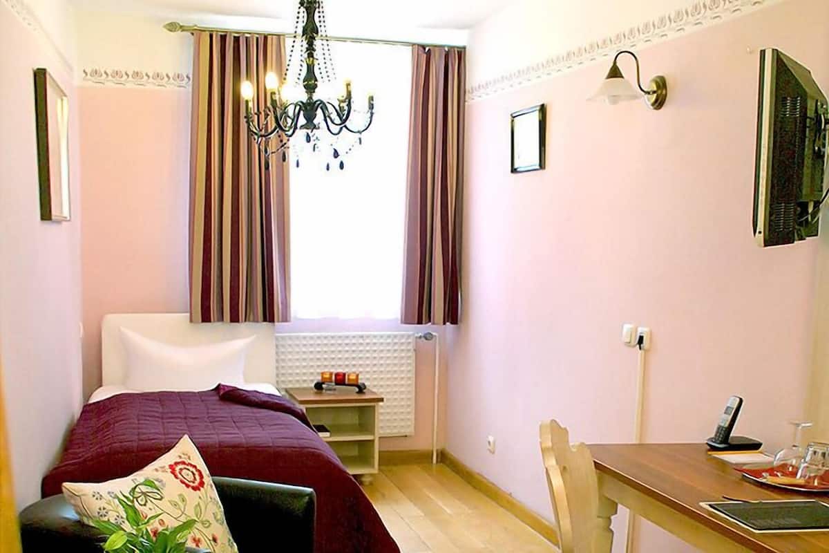 A hotel room with a bed, a desk, a chair, a chandelier, a window with curtains, a radiator, a wall-mounted TV, and a bedside table with a lamp and a phone.