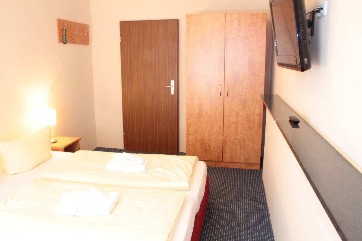 Ein Hotelzimmer mit Bett, Kleiderschrank, Wandfernseher und einem kleinen Tisch mit Lampe.