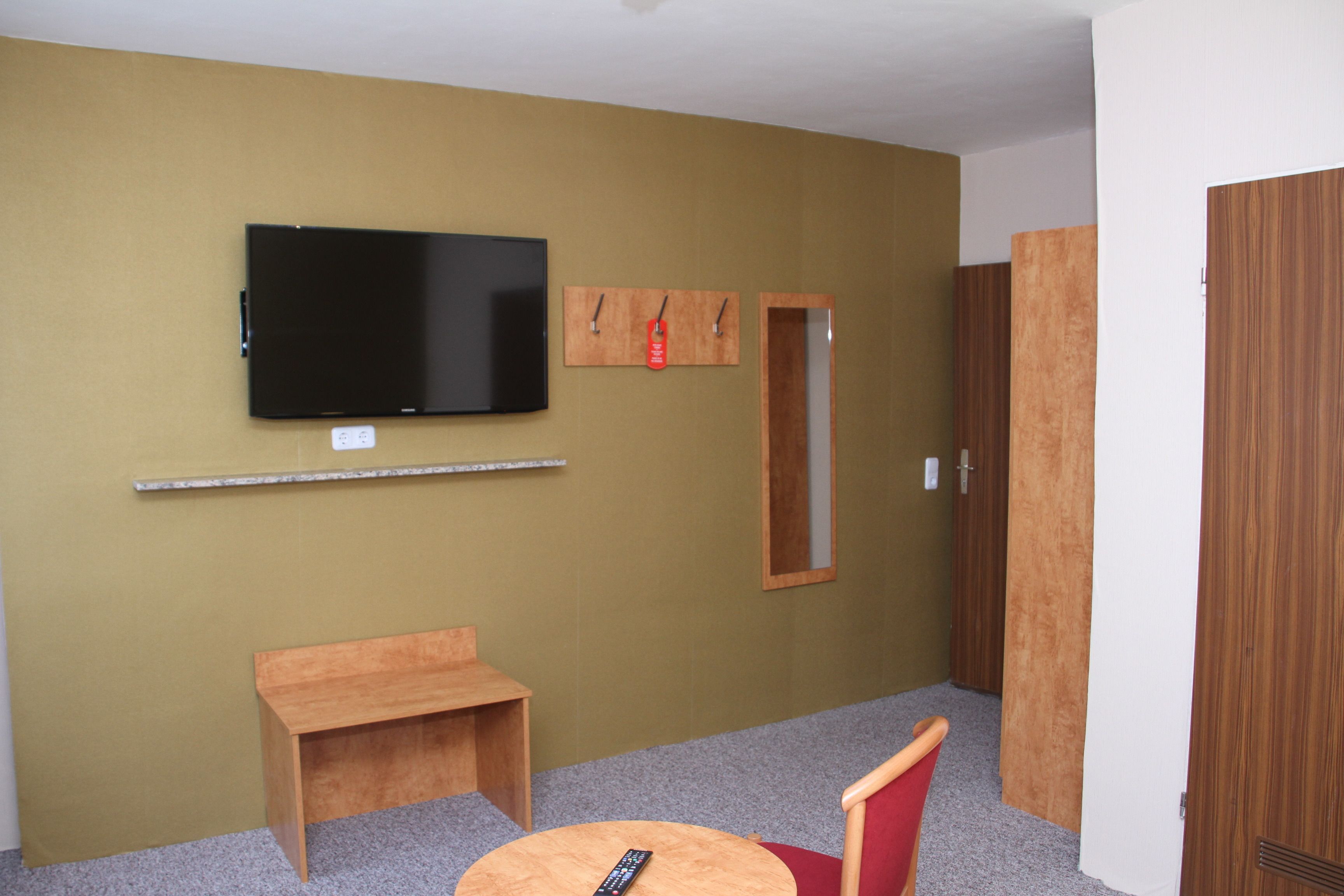 A room with a flat-screen TV mounted on the wall, a small wooden desk, a red chair, and a wooden wardrobe.