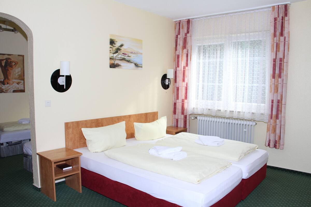 Ein Hotelzimmer mit einem Doppelbett, einem Nachttisch aus Holz, einem Heizkörper und einem Fenster mit Vorhängen.