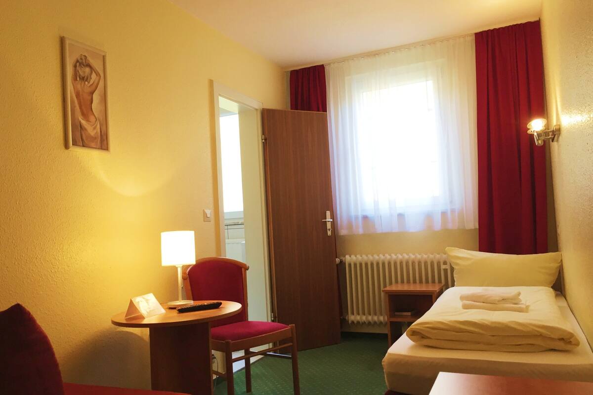 Ein Hotelzimmer mit Bett, Schreibtisch mit Stuhl, Lampe, einem Bild an der Wand und einem Fenster mit Vorhängen.