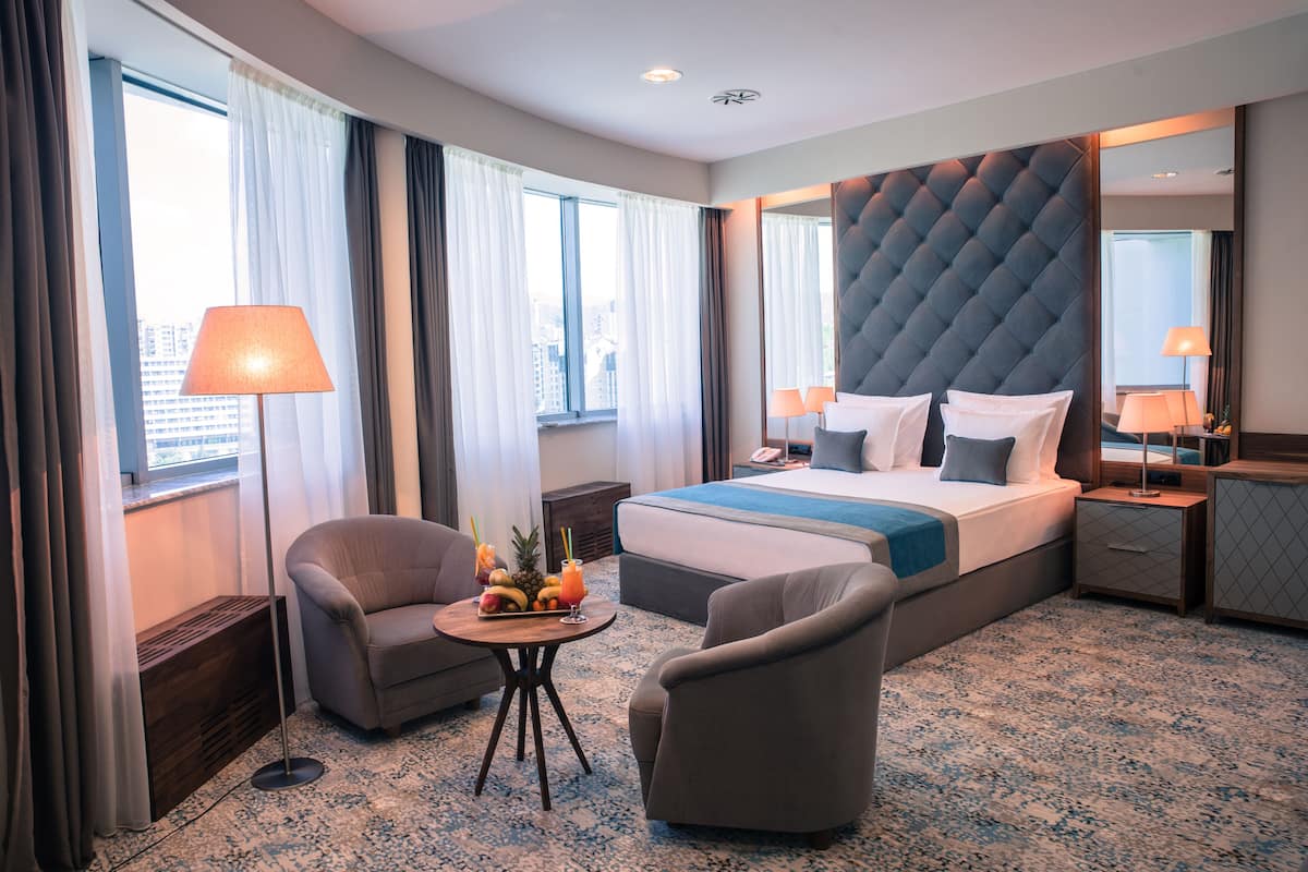Habitación de hotel con una cama grande, dos sillones, una mesita con fruta y vistas a la ciudad a través de amplios ventanales.