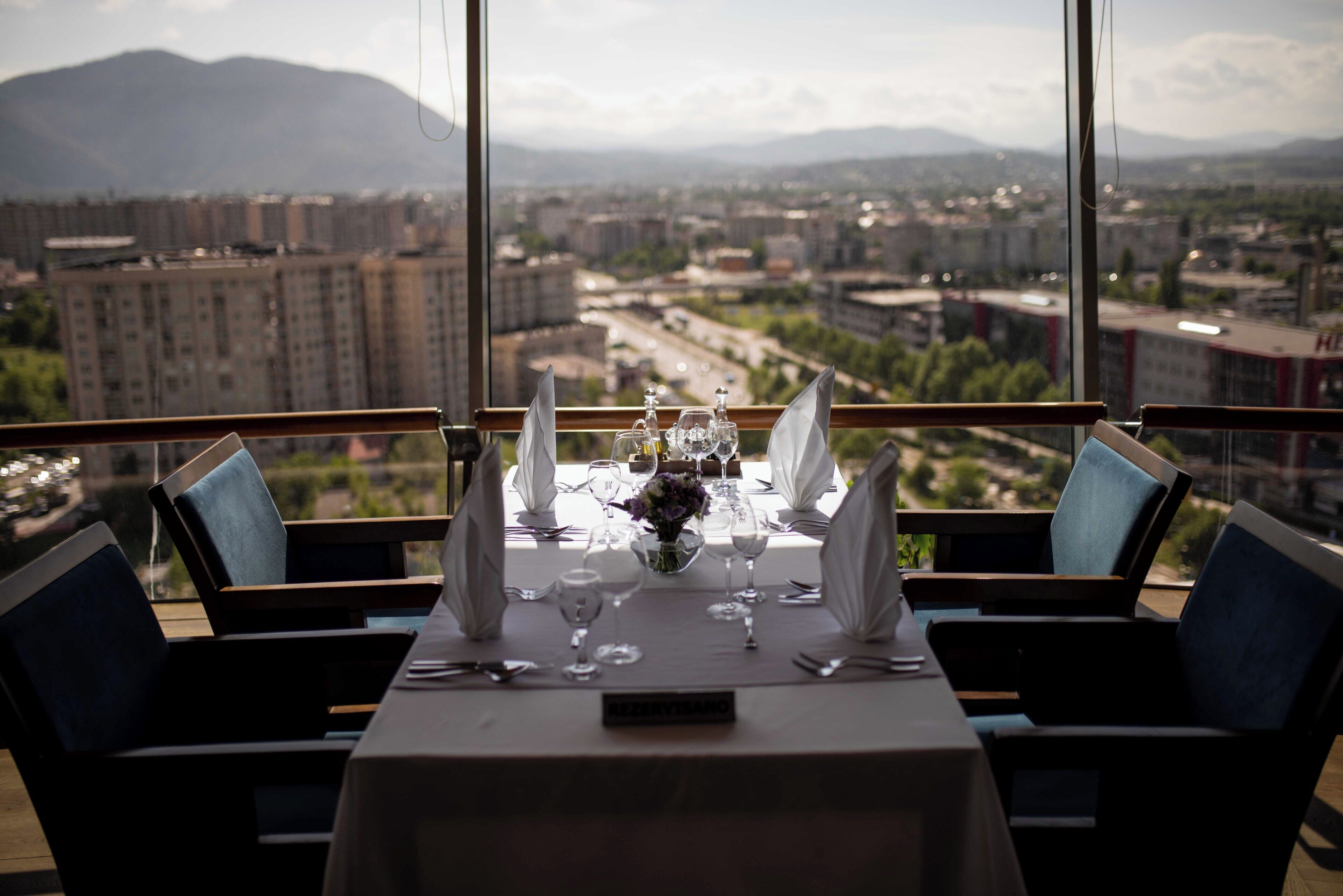 A dining table set for two with a city view, chairs, and a vase with flowers.