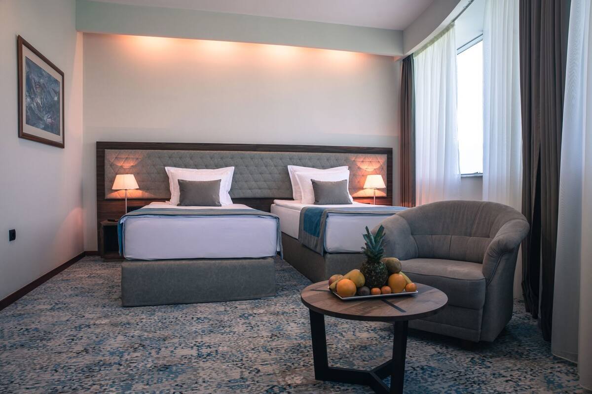 Una habitación de hotel con dos camas, un sillón gris, una mesa redonda con fruta y una lámpara.