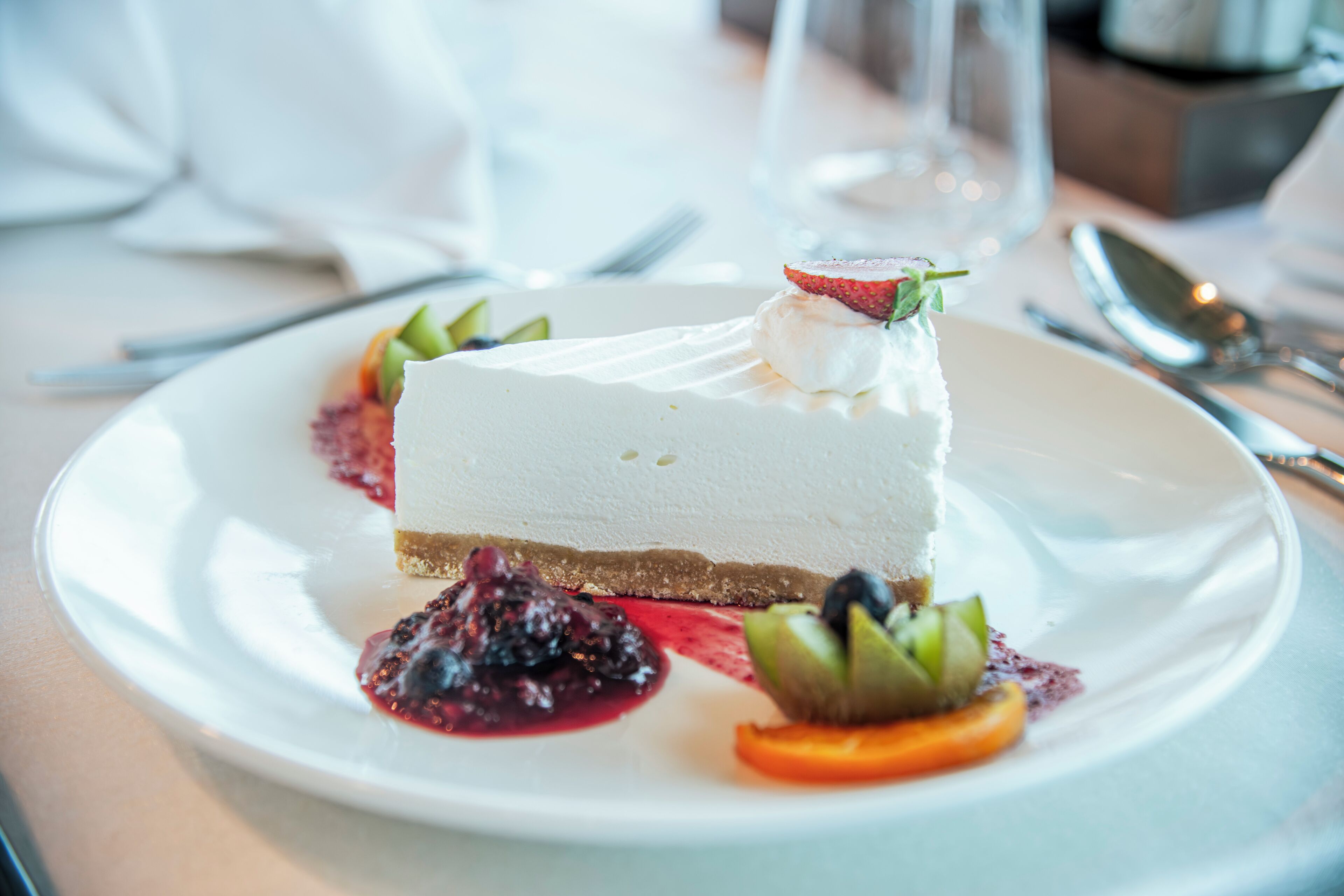 Una porción de pastel de queso sobre un plato blanco, con coulis de frutos rojos y decoración de frutas.