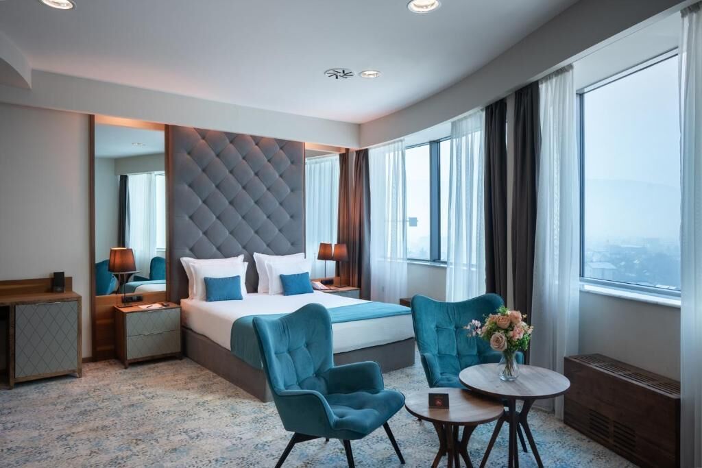 Ett hotellrum med en stor säng, en sittgrupp med bord och stolar, en spegel och utsikt över staden.