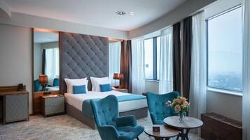 Ett hotellrum med en stor säng, en sittgrupp med bord och stolar, en spegel och utsikt över staden.