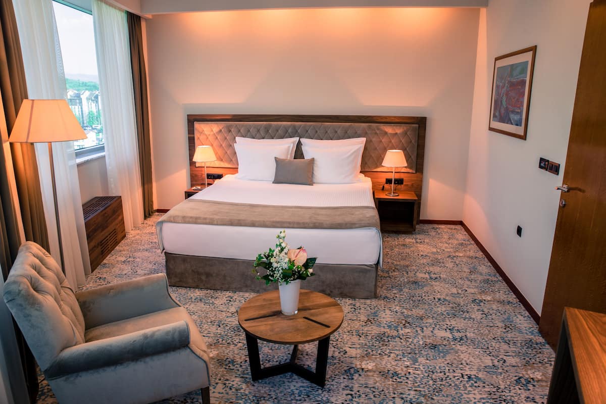 Habitación de hotel con una cama grande, una mesita de noche, una silla, una pequeña mesa redonda con un florero y una ventana con cortinas.