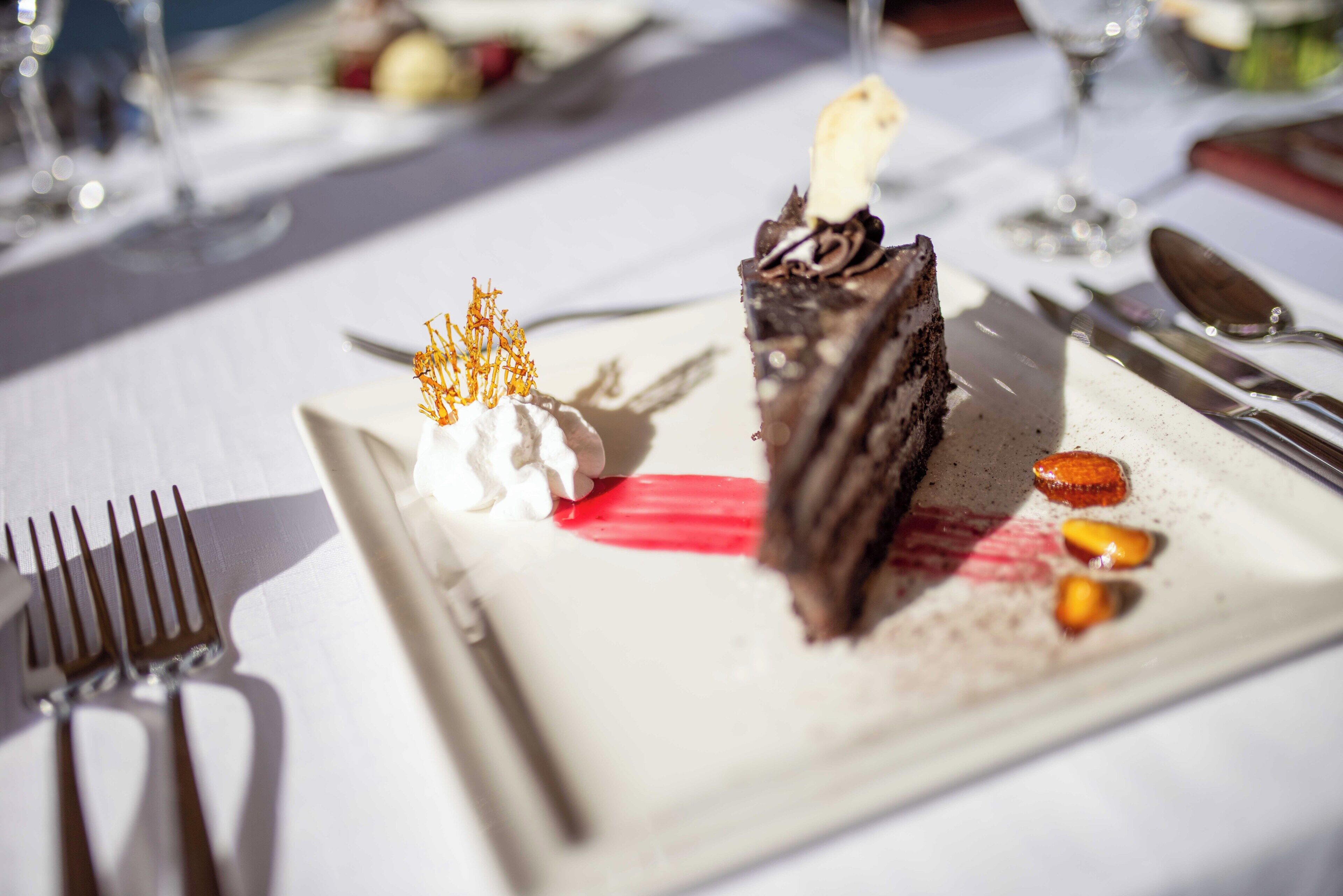 Un plat de postre amb una porció de pastís, nata muntada i una espelma decorativa sobre una taula amb coberts.