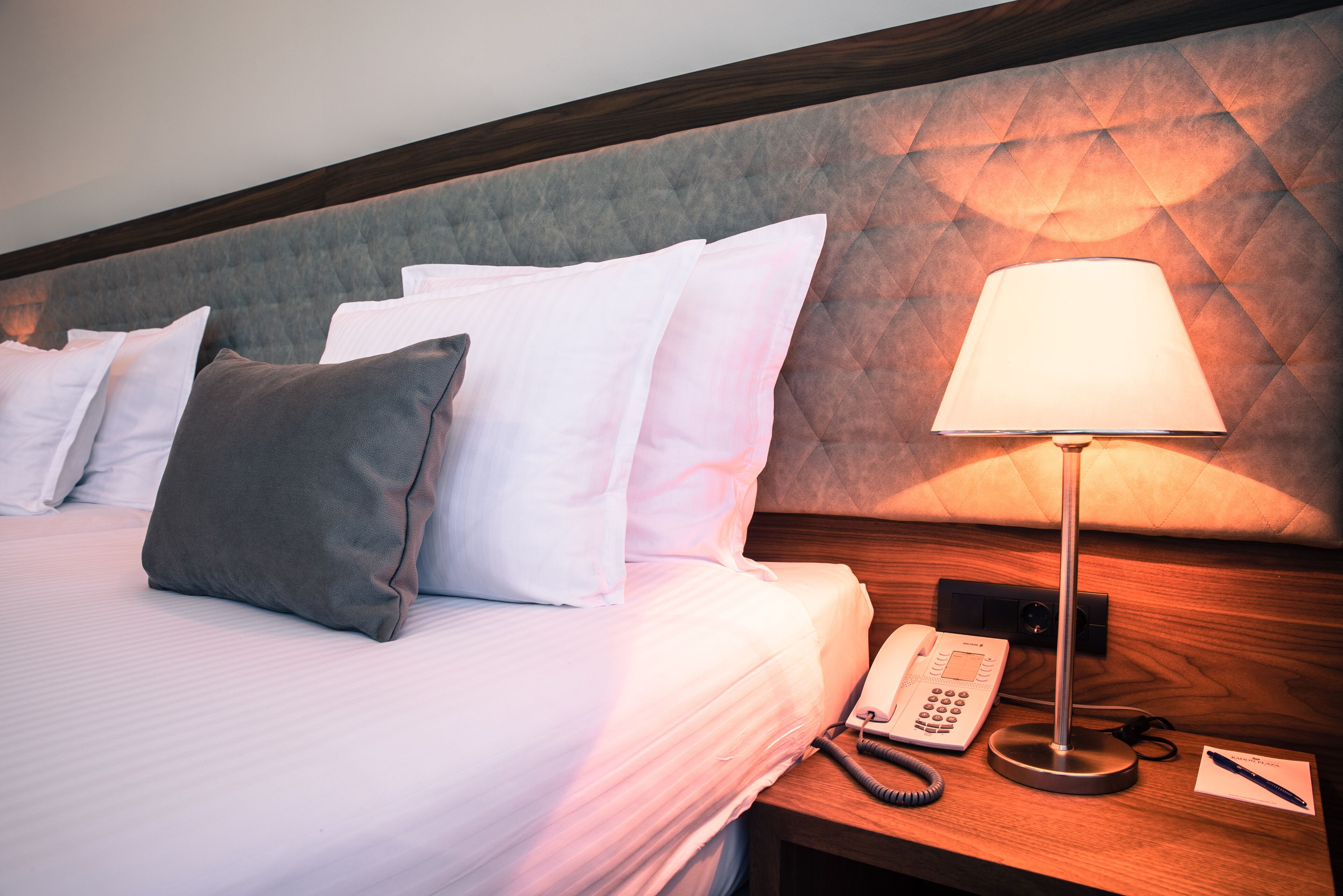A hotel room with a bed, pillows, a bedside lamp, a phone, and a notepad.