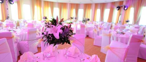A banquet hall with round tables, white chairs, and floral centerpieces.