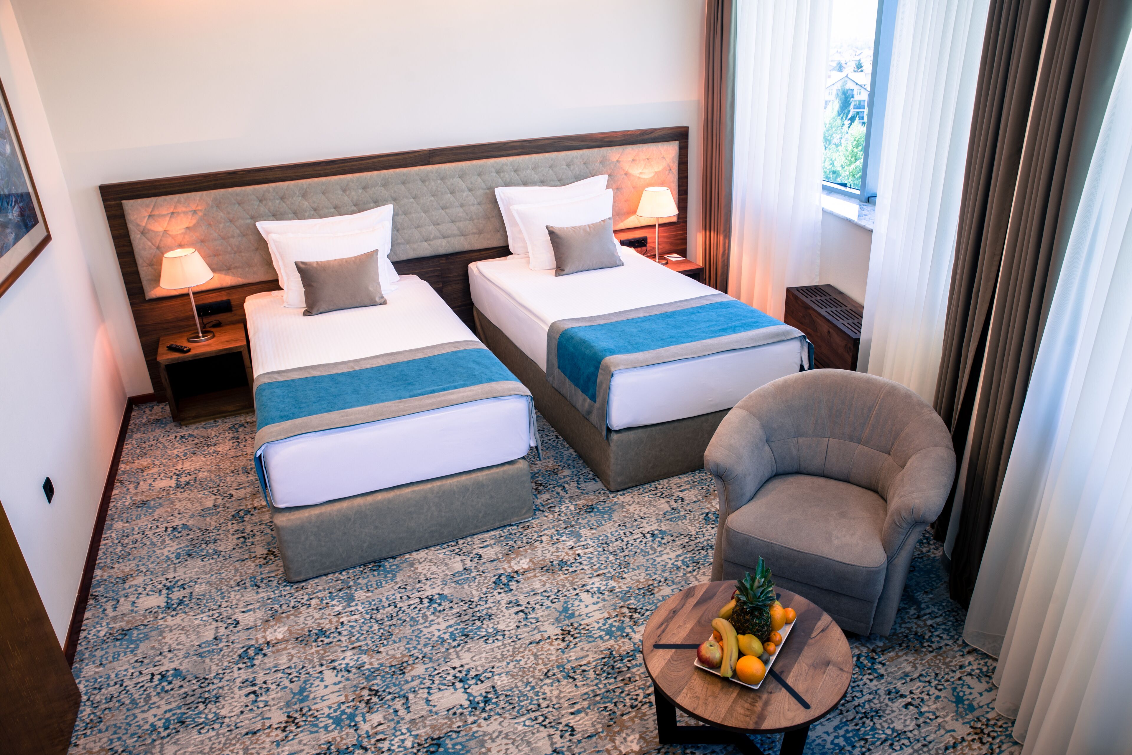 A hotel room with two beds, a small armchair, a round wooden table with fruit, and a view of greenery through the window.