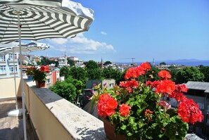 A balcony with red geraniums, a striped umbrella, and a view of a cityscape with buildings and a distant sea. - Fors Hotel Sultanahmet (Istanbul)