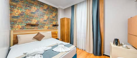 A hotel room with a bed, a wooden wardrobe, a bedside table with a kettle, and a large window with curtains.