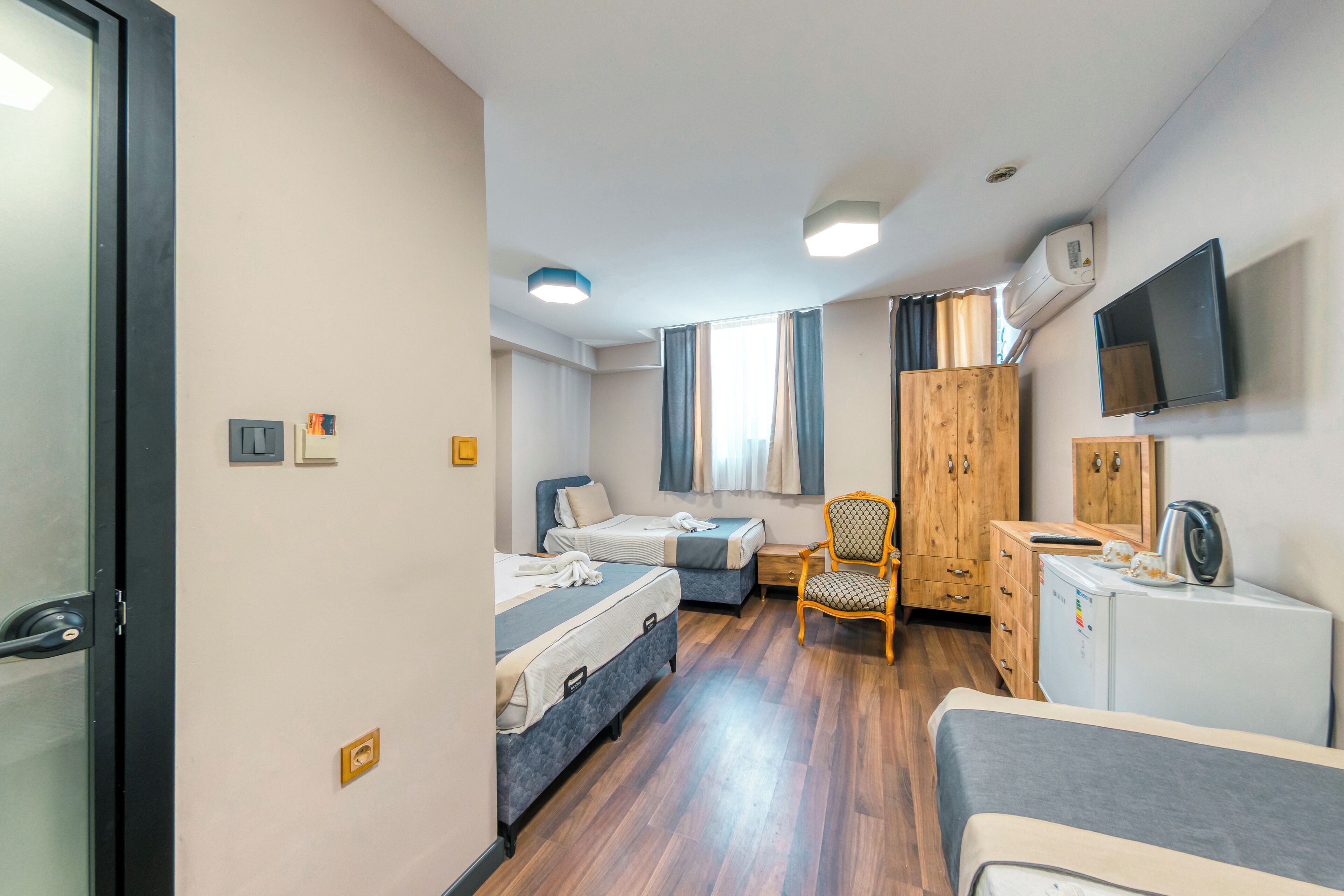 A hotel room with two beds, a wooden wardrobe, a desk with a kettle, a chair, and a TV mounted on the wall.