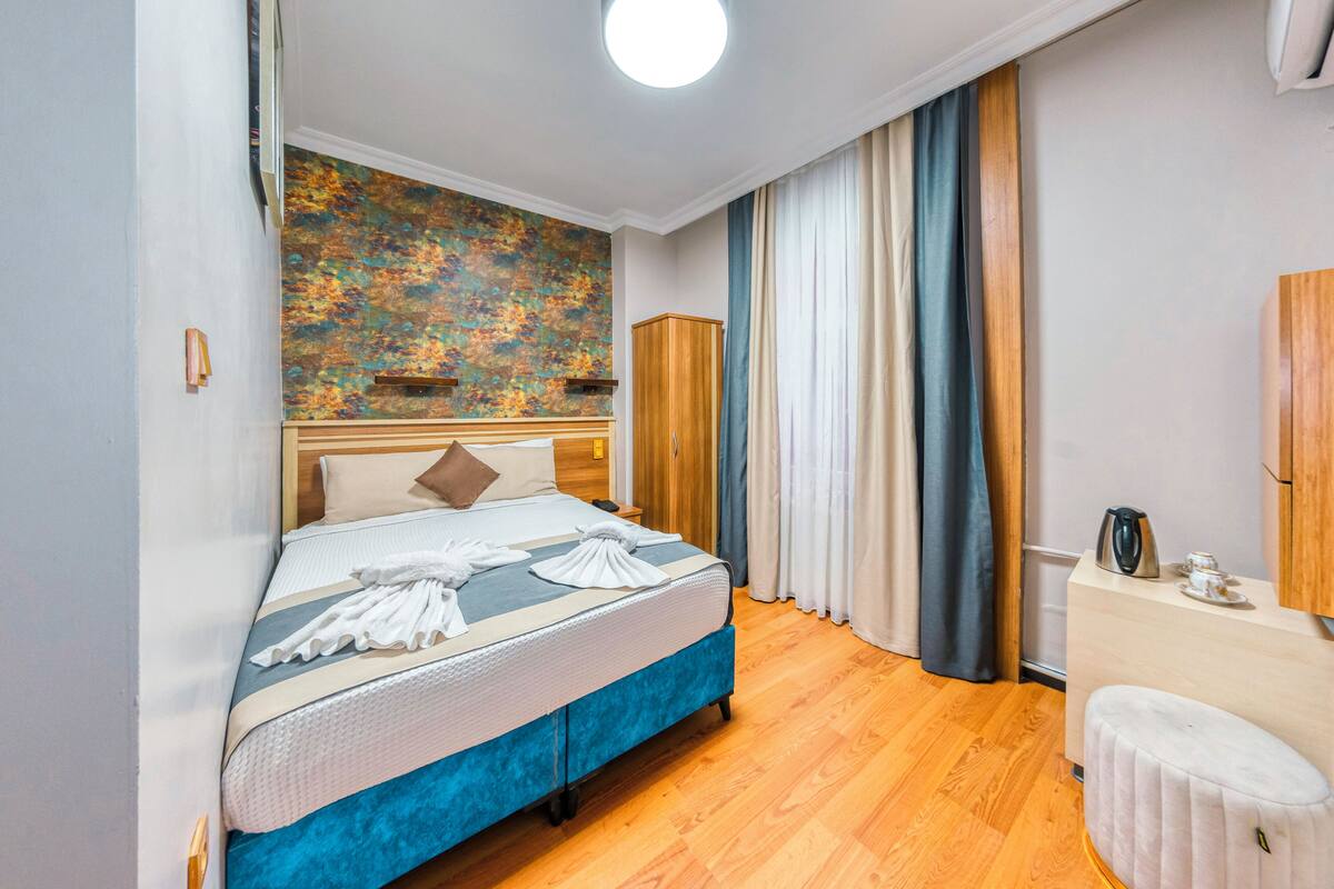 A neatly arranged bedroom with a bed, a wooden wardrobe, a small table with a kettle, and a wall with a textured artwork.