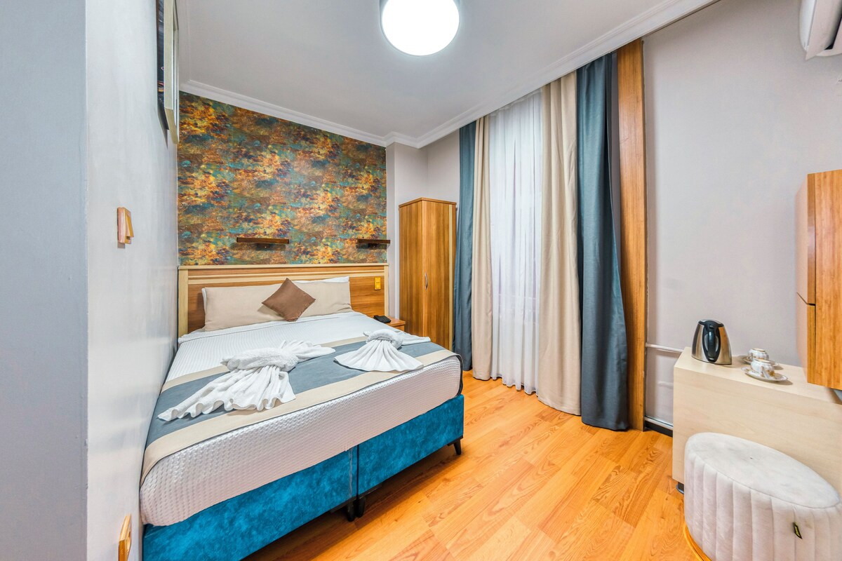 A neatly arranged bedroom with a bed, a wooden wardrobe, a small table with a kettle, and a wall with a textured artwork.