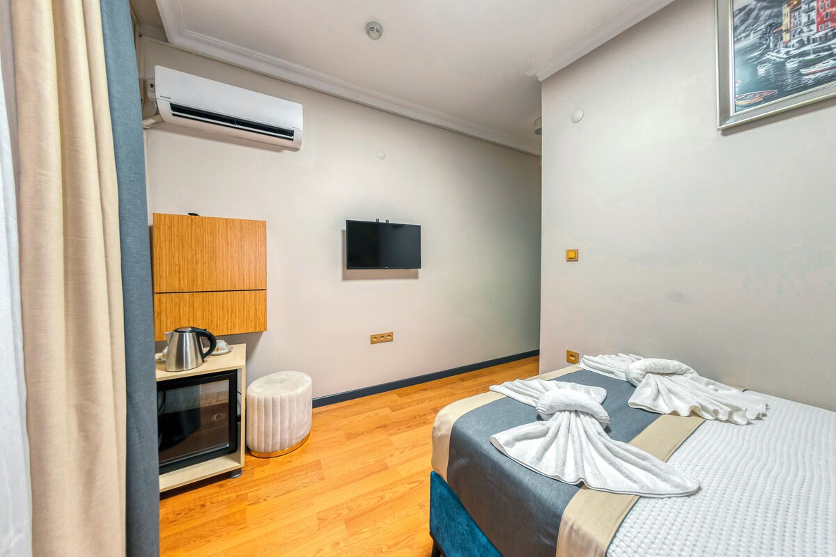 A hotel room with a bed, a TV, an air conditioner, a small kitchenette, and a wooden cabinet.
