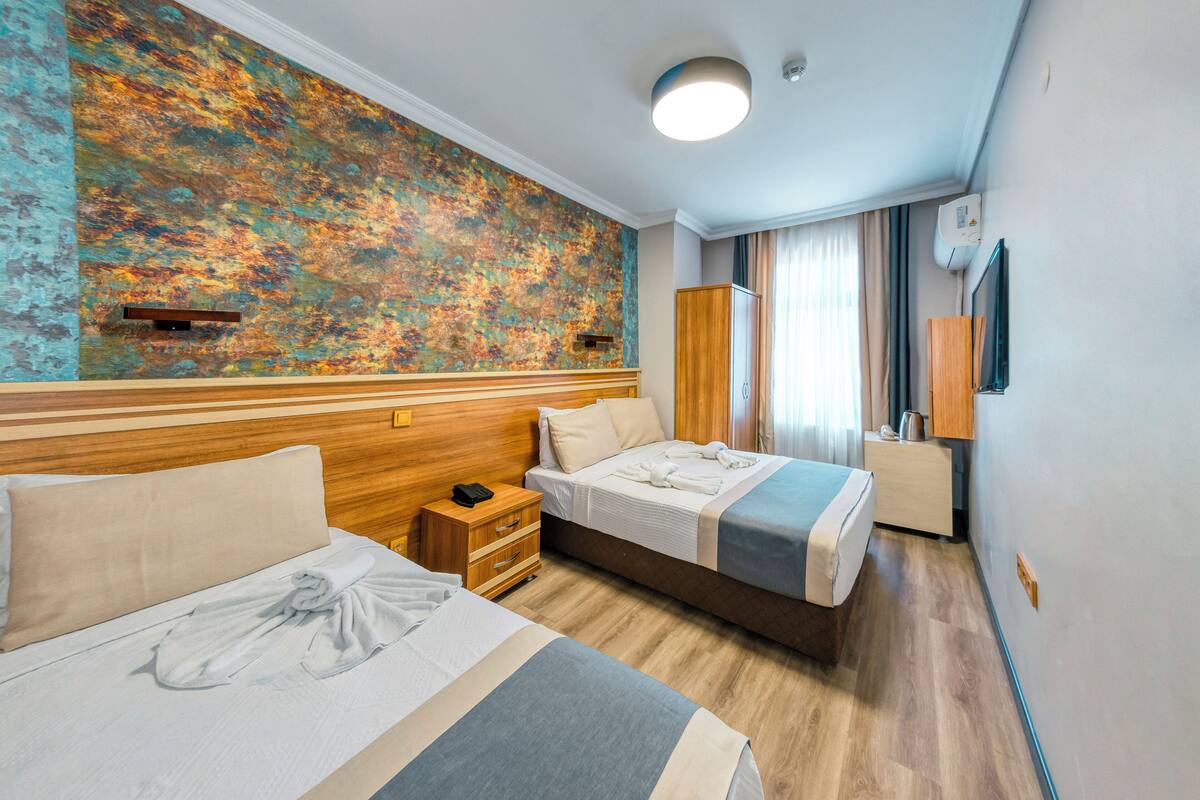 A hotel room with two beds, a wooden headboard, a large abstract mural, a wooden nightstand, a wardrobe, and a window with curtains.