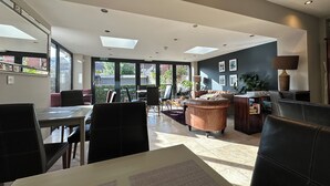 Ein großzügiger Wohnbereich mit Esstisch, Stühlen, einer Couch und einem Couchtisch. Große Fenster sorgen für natürliches Licht und bieten Ausblick ins Freie.