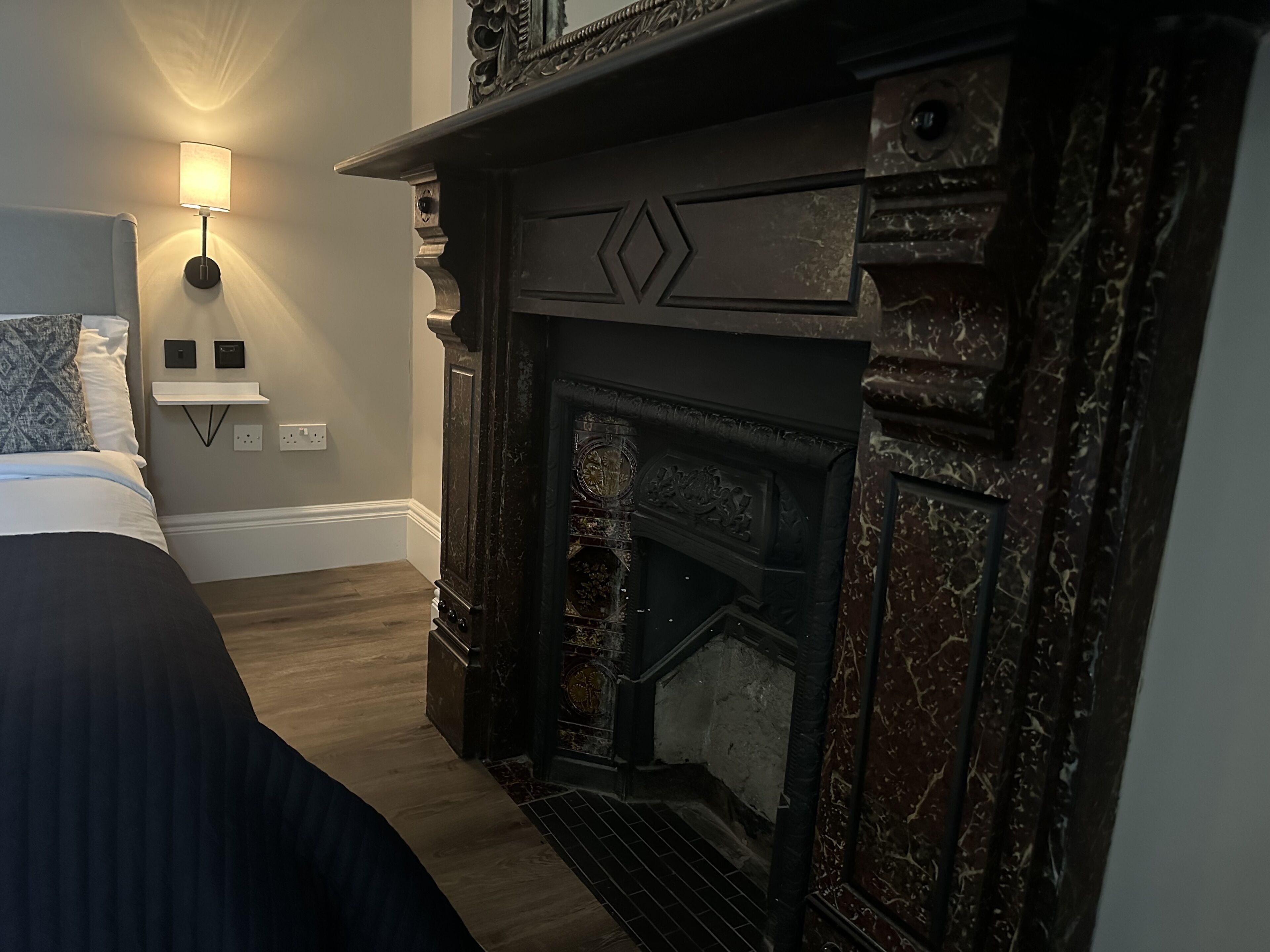 A bedroom with a bed, a wall-mounted lamp, a fireplace, and a small shelf.
