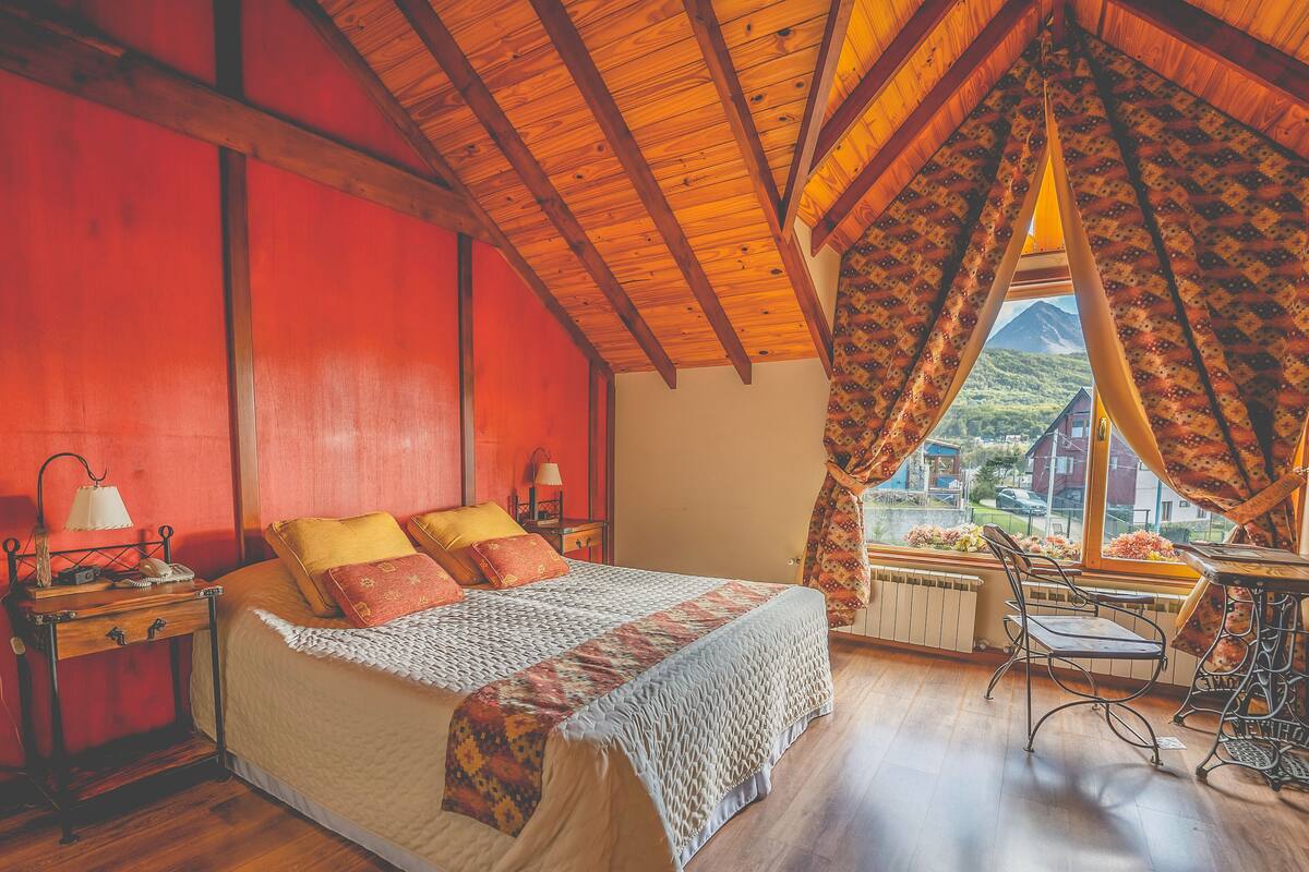 Ein Schlafzimmer mit Holzbalkendecke, einem Bett mit gemusterten Bettwaren, einem Nachttisch mit Lampe und Blick ins Grüne.