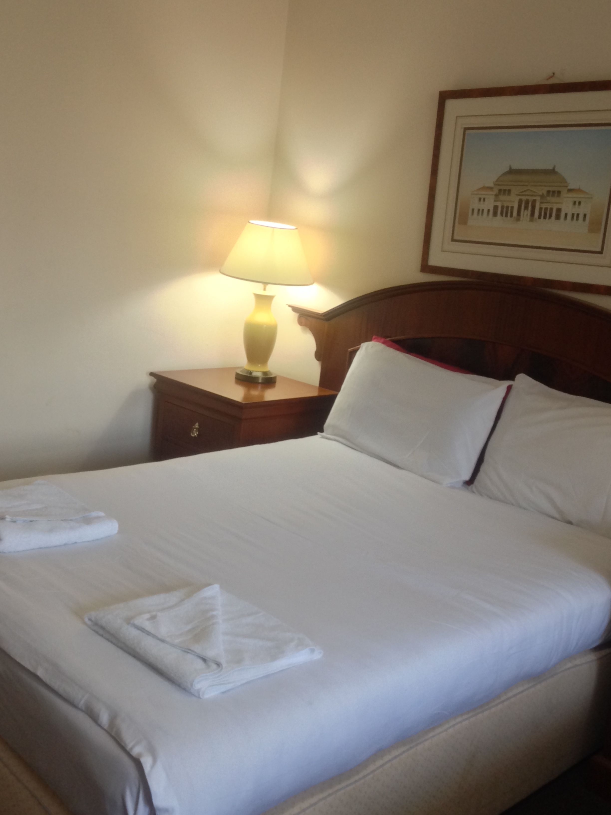 A neatly made bed with white linens and a folded towel, a wooden nightstand with a lamp, and a framed picture on the wall.