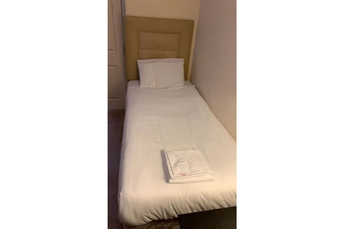 Ein Einzelbett mit weißer Matratze und Kopfteil, ein gefaltetes weißes Handtuch auf dem Bett und eine schlichte Wand im Hintergrund.
