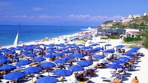 A beach with numerous blue umbrellas and sun loungers, a clear blue sea, and buildings in the background. - LABRANDA Rocca Nettuno Tropea (Tropea)
