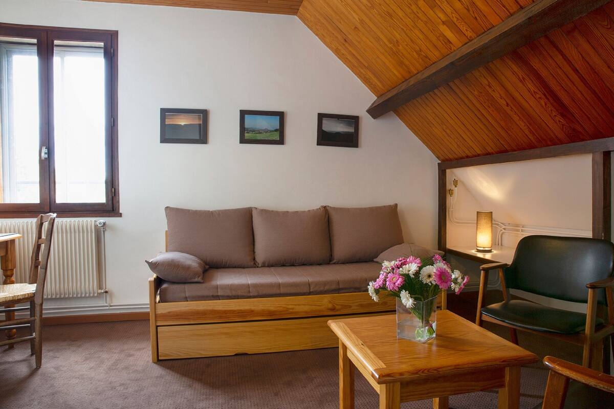 Un salon chaleureux avec un canapé marron, une table basse en bois et un vase rempli de fleurs. Trois tableaux encadrés ornent le mur, et une fenêtre offre une vue sur l’extérieur.