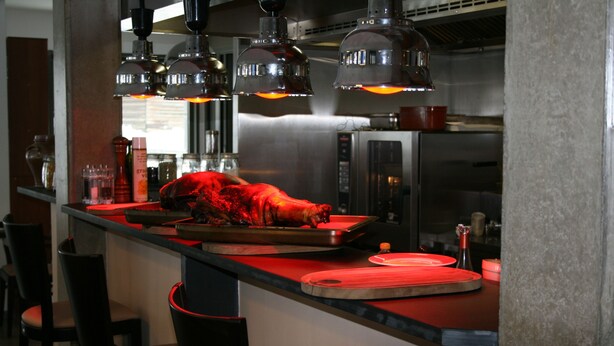 A kitchen with a large, red-roasted meat on a tray, overhead industrial-style lighting, and stainless steel appliances. - The Originals Relais, Auberge de la Petite Ferme (Besse-et-Saint-Anastaise)