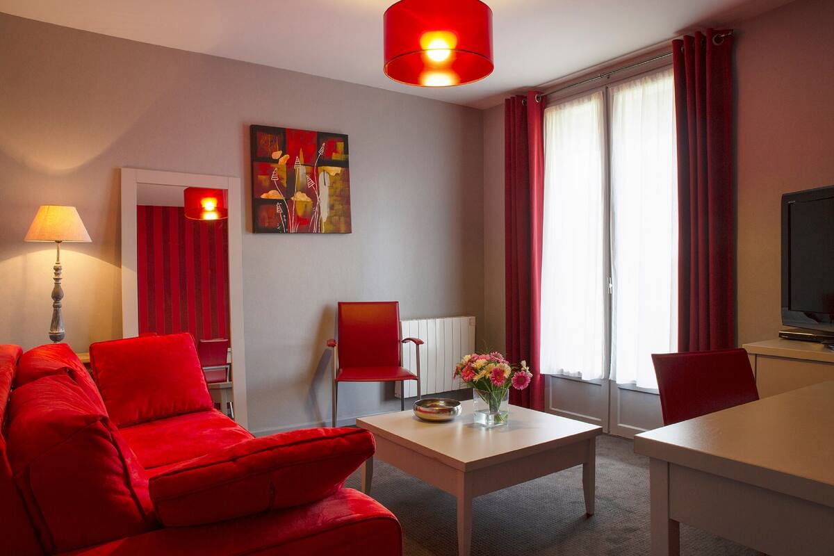 Une chambre d’hôtel moderne avec un canapé rouge, une table à manger, un téléviseur et un tableau au mur.