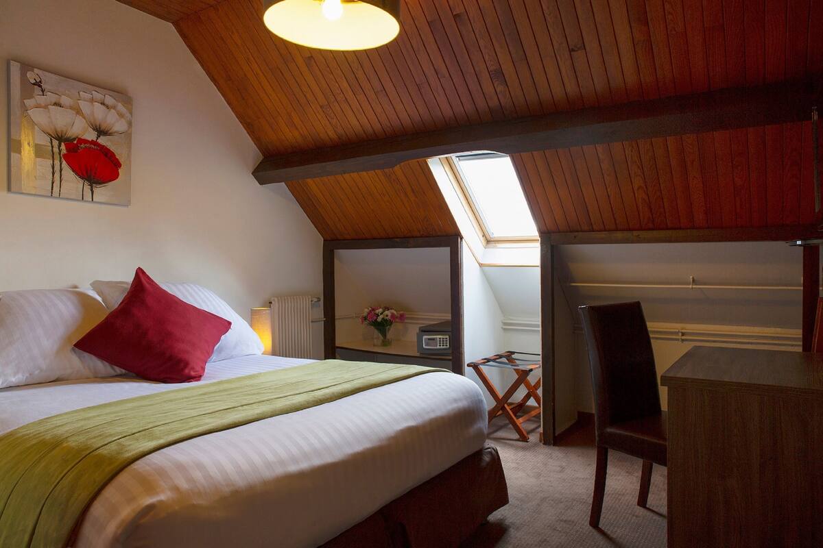 Une chambre à coucher avec un lit, un oreiller rouge, un plaid vert, un bureau en bois et une chaise.