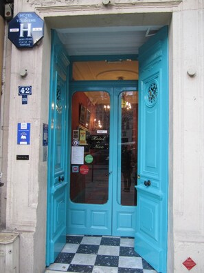 A hotel entrance with a turquoise door, a sign indicating it is a tourist hotel, and a check-in desk visible through the glass. - Hotel De Nice (Paris)