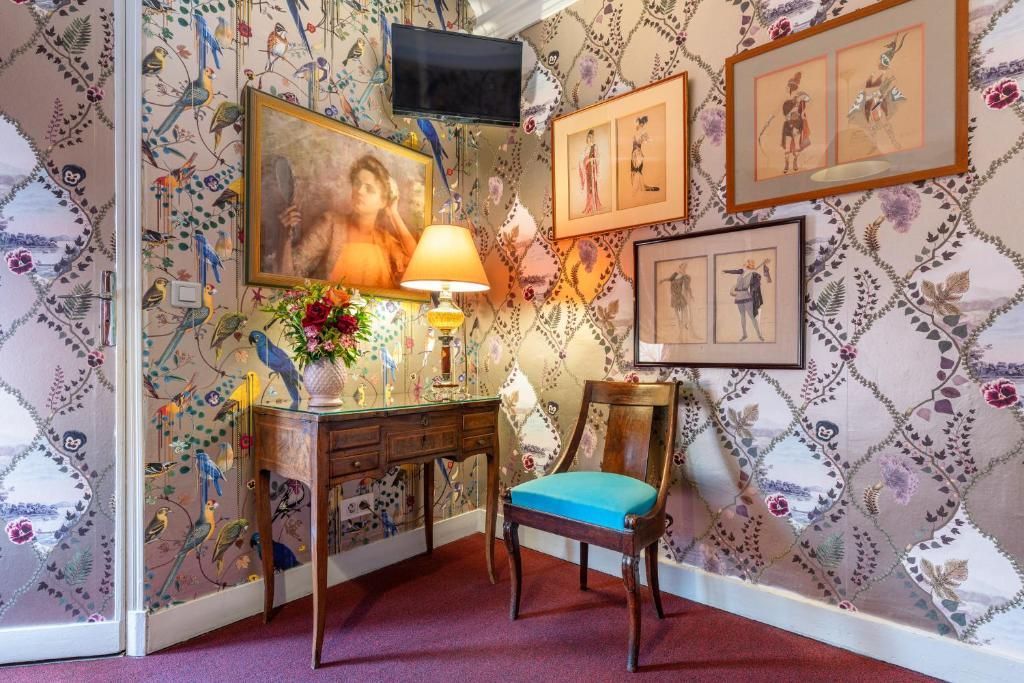 A room with a patterned wallpaper, a desk with a lamp, a chair, and framed pictures on the wall.