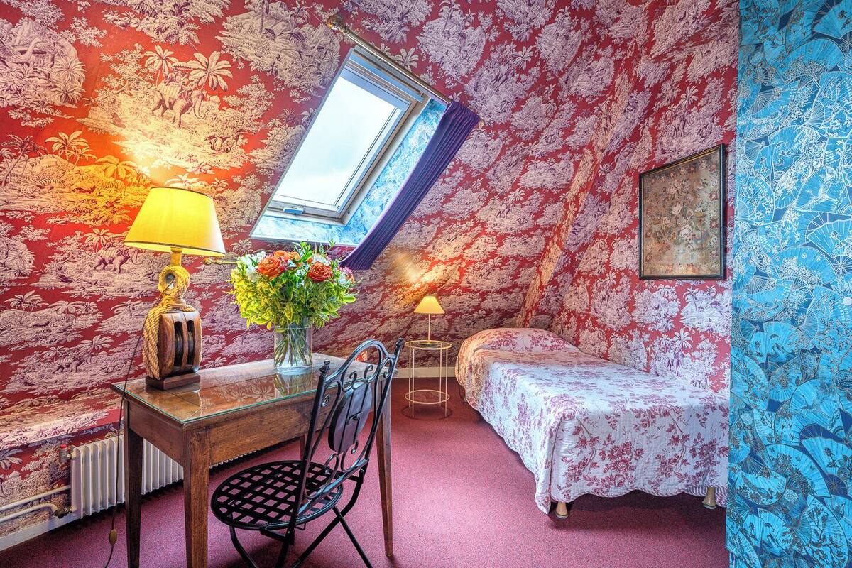A bedroom with a sloped ceiling, floral wallpaper, a skylight, a wooden table with a lamp, a chair, a bed with a floral bedspread, and a small bedside table with a lamp.