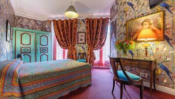 Una habitación con cama, escritorio con lámpara y silla. Hay una ventana con cortinas, un mural de pájaros en la pared y un cuadro en la pared.