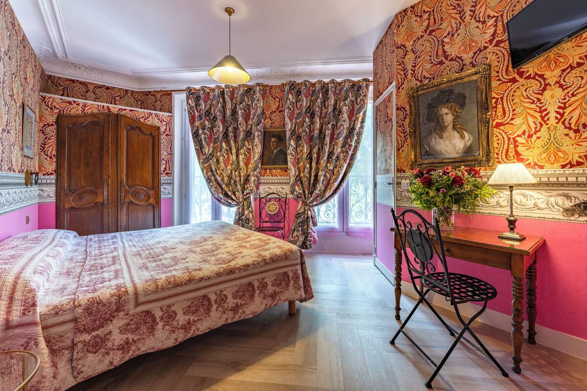 A room with a bed, a desk with a chair, a lamp, a painting on the wall, and curtains.