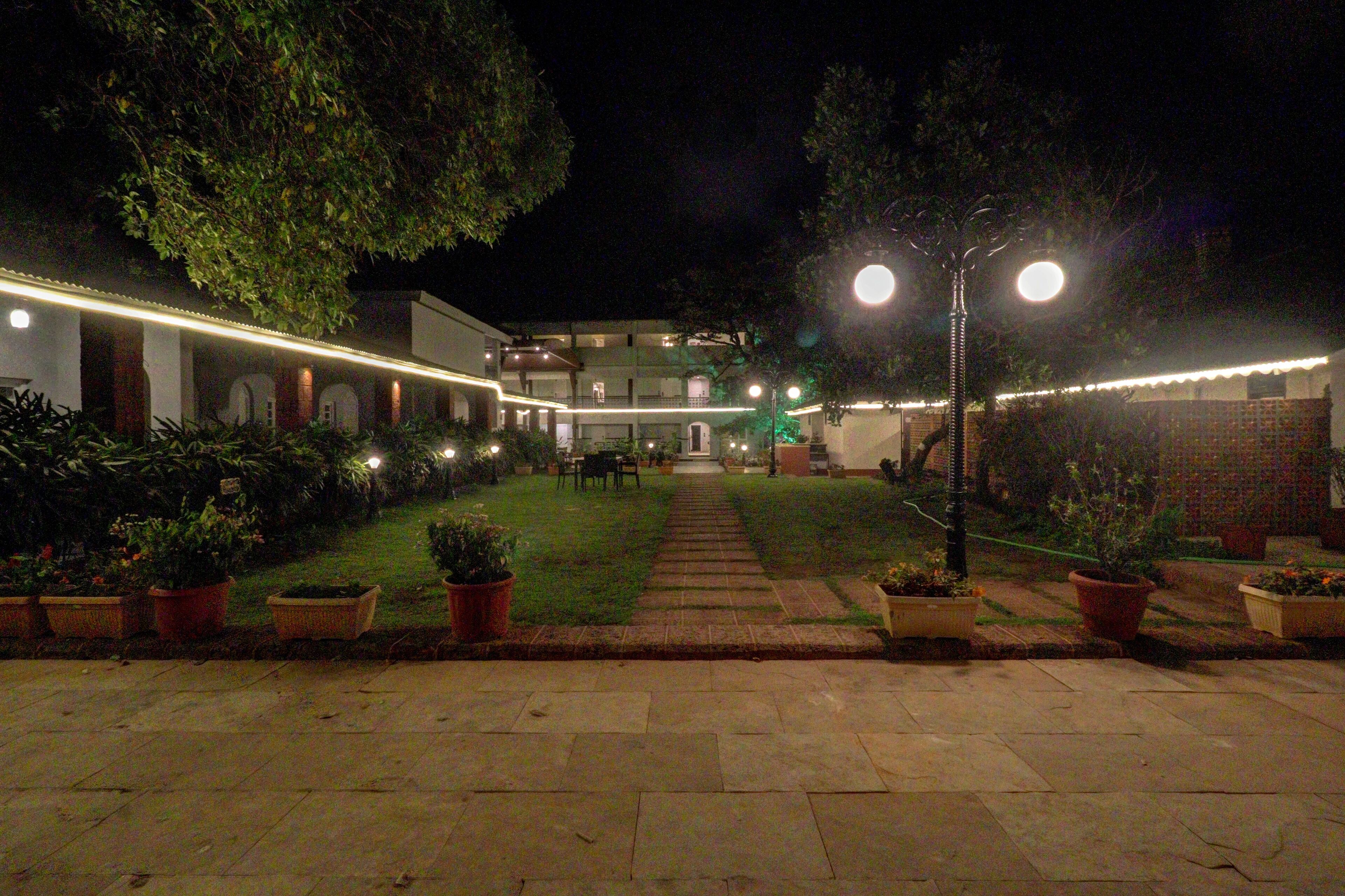 A courtyard with a pathway leading to a building, surrounded by potted plants and illuminated by street lamps.