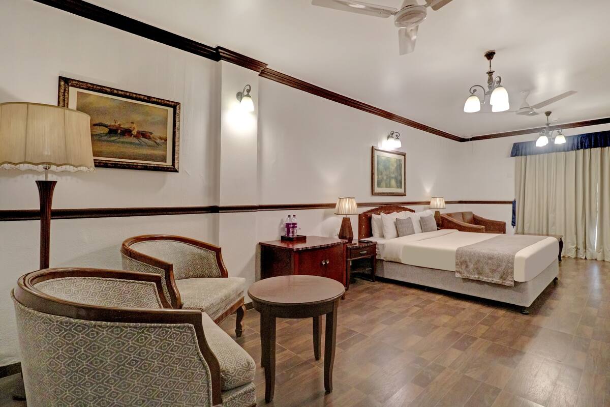 A hotel room with a bed, two chairs, a small table, a nightstand, and a painting on the wall.