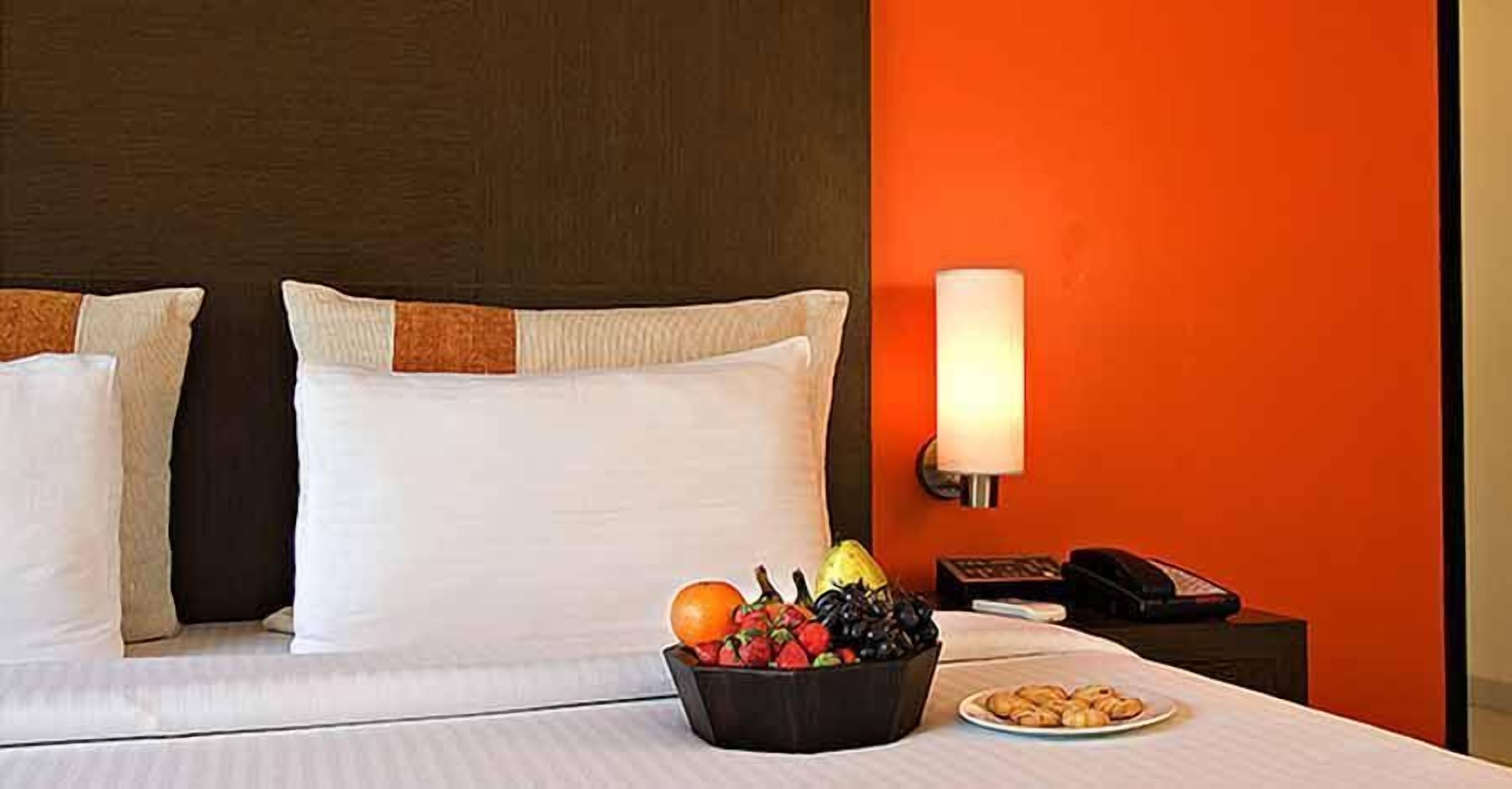 A hotel room with a bed, a fruit bowl, a plate of cookies, a bedside lamp, and a telephone on a nightstand.