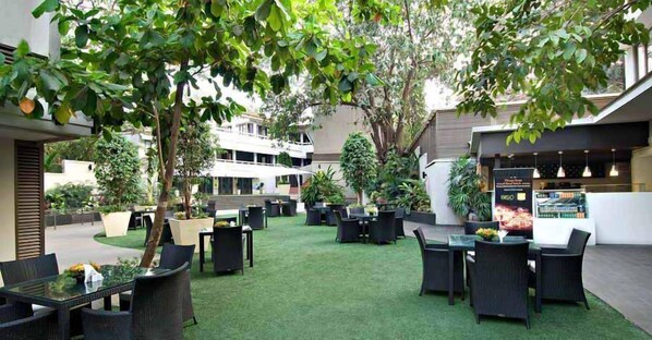 Relax in a comfortable outdoor space with tables, chairs, and lush artificial grass, surrounded by trees and shrubs.