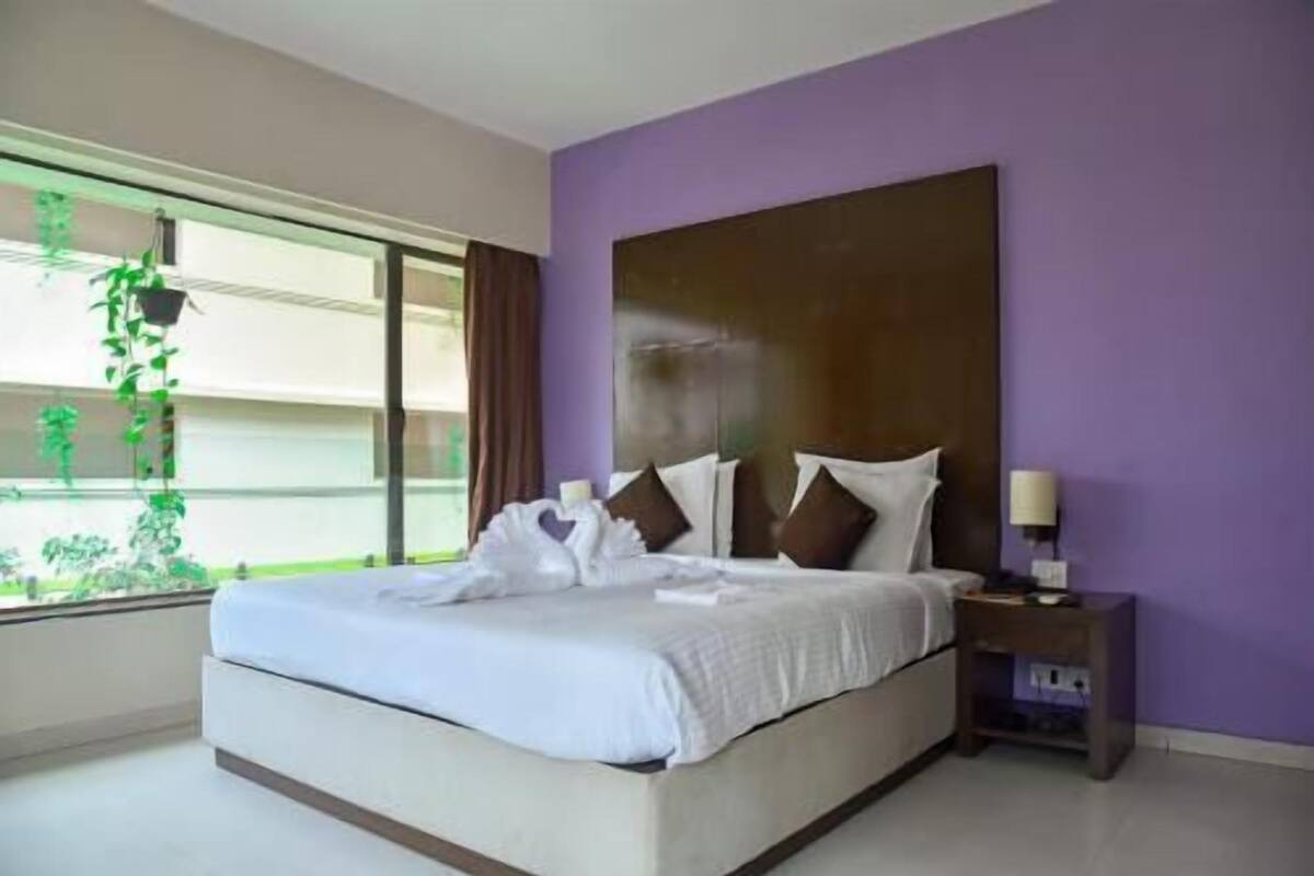 A bedroom with a bed, a nightstand, a window with plants, and a purple wall.