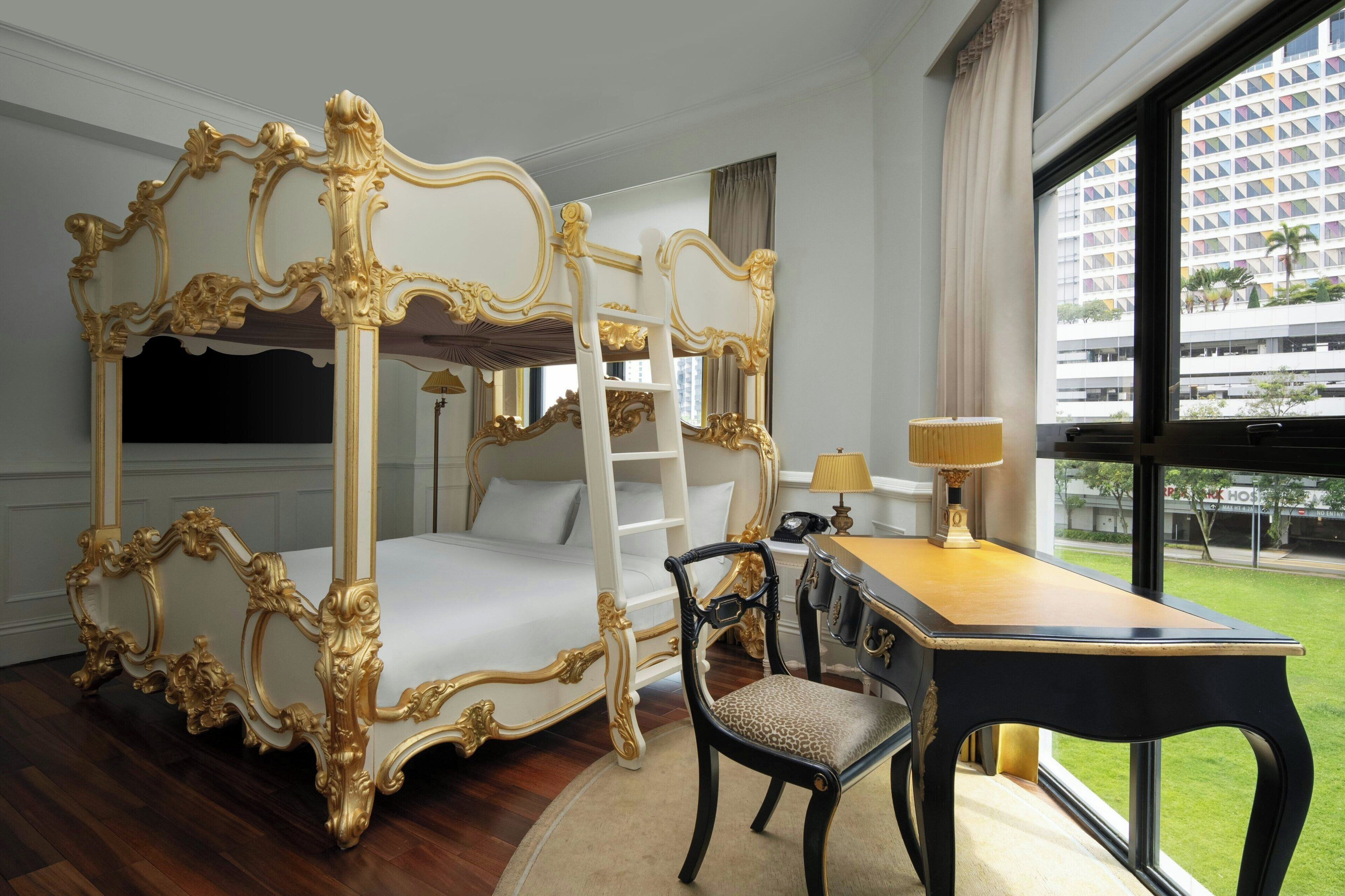 Una cama con dosel y detalles dorados ornamentales, un escritorio con lámpara y vistas a un edificio a través de un ventanal grande.