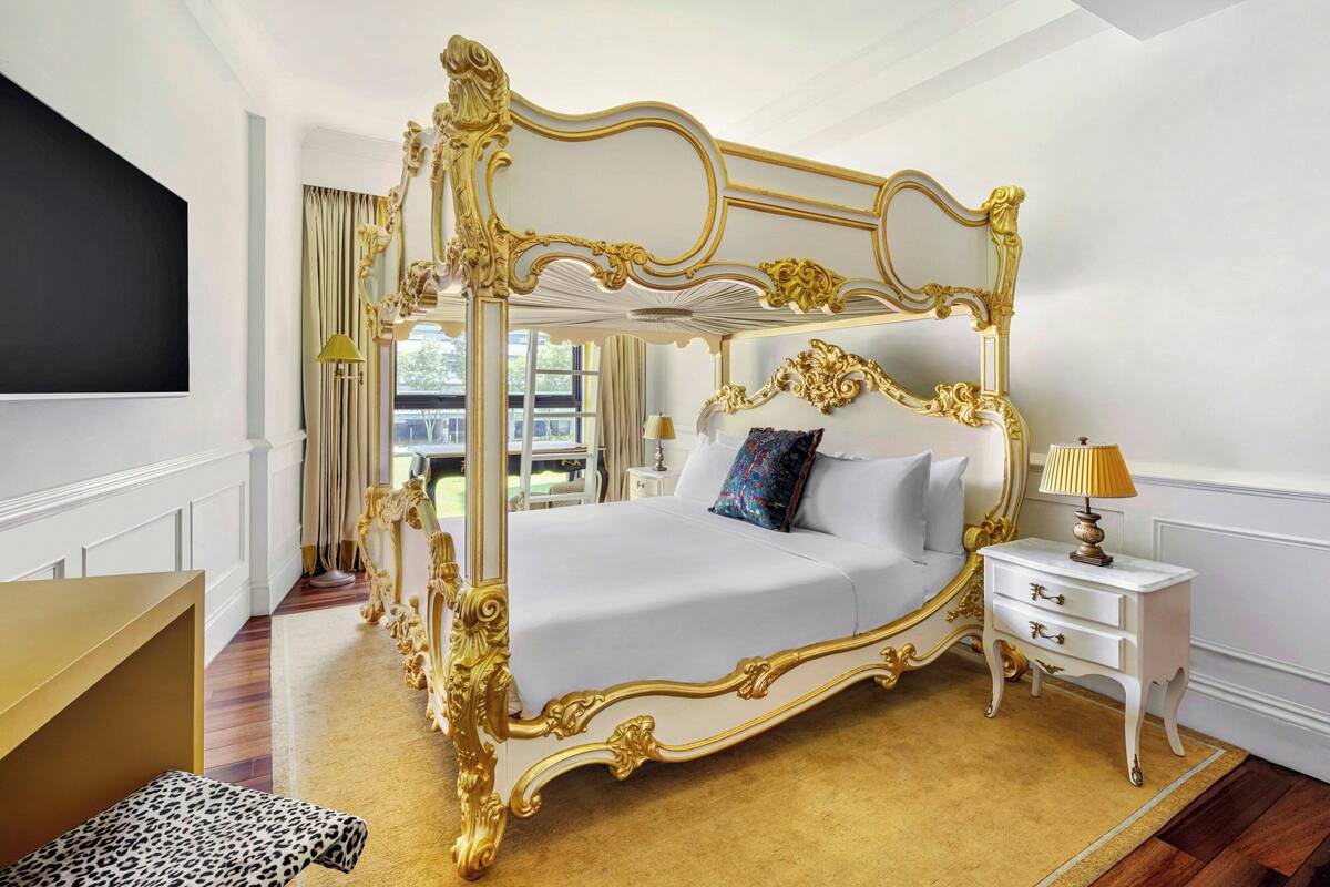 Un letto a baldacchino con dettagli dorati decorativi, una testiera imbottita bianca e un comodino abbinato con cassetti.